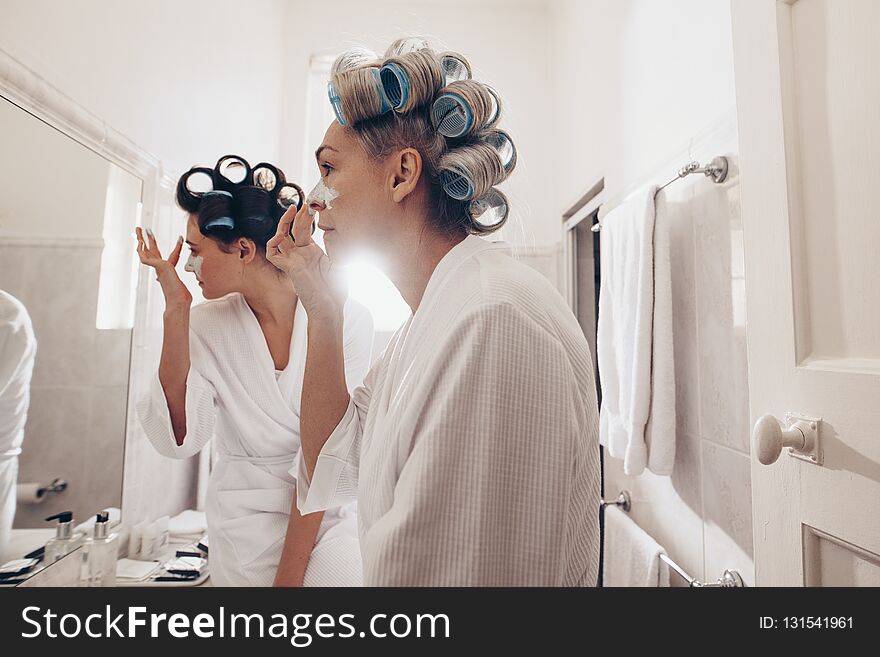 Side view of two women in bathrobes applying cream on face standing in front of mirror. Women with curly rollers pinned on hair