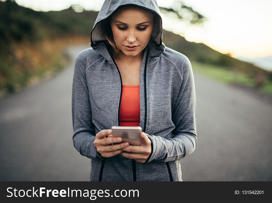 Fitness woman walking on street wearing a hooded sweat shirt looking at her mobile phone. Woman using her cell phone walking on an