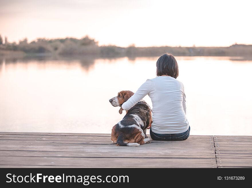 Pretty young woman hugging her dog Basset Hound on the wooden dock on the river