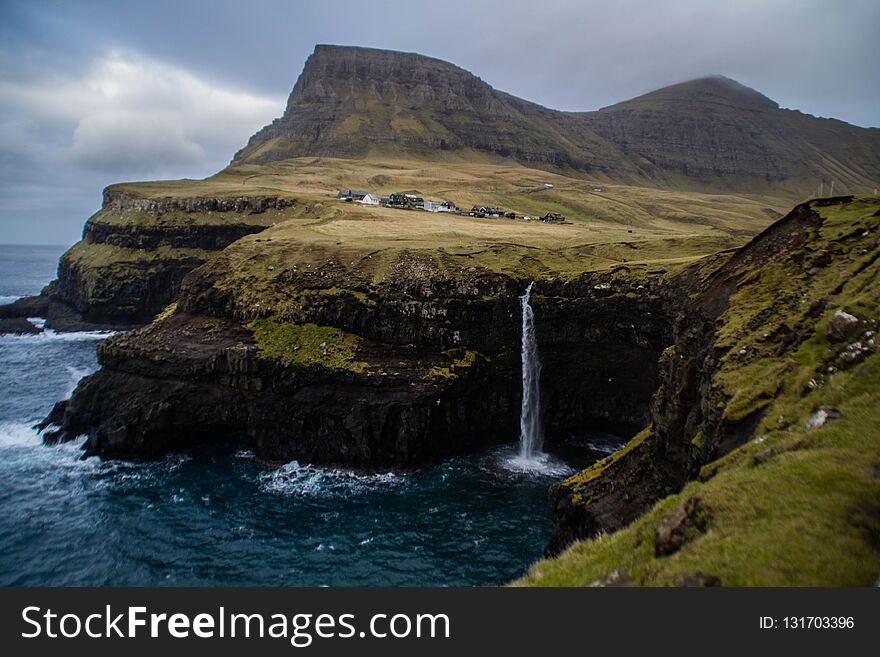 Travel/Sightseeing Faroe IslandsDenmark. Scenic panorama landscape view of cliffs, waterfall and Gasadalur village on background. Tourist popular spot/place in VÃ¡gar Island. Moody dramatic sky. Travel/Sightseeing Faroe IslandsDenmark. Scenic panorama landscape view of cliffs, waterfall and Gasadalur village on background. Tourist popular spot/place in VÃ¡gar Island. Moody dramatic sky.