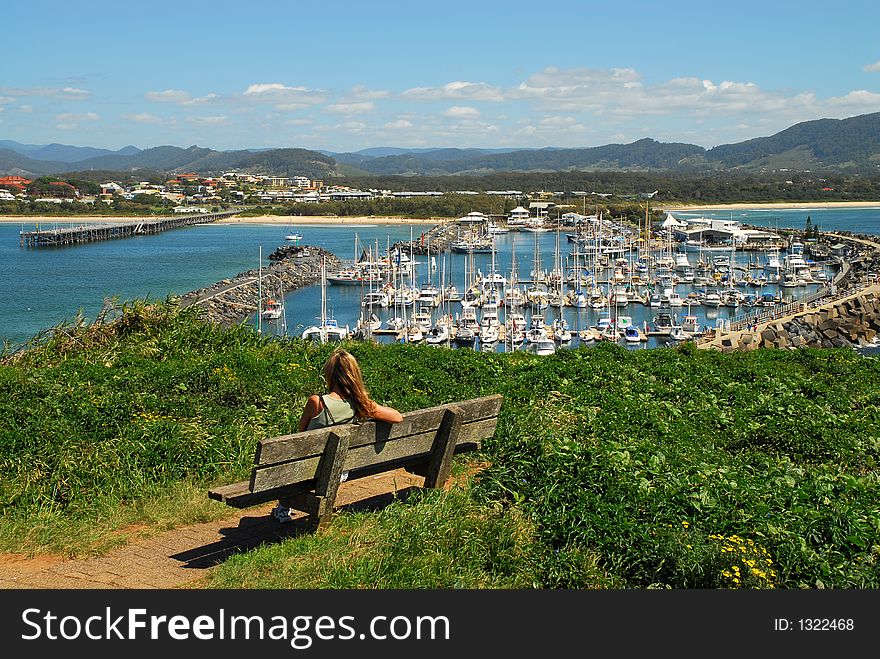 A girl seated on a bench looking down at the marina with sailing boats moored there. A girl seated on a bench looking down at the marina with sailing boats moored there.