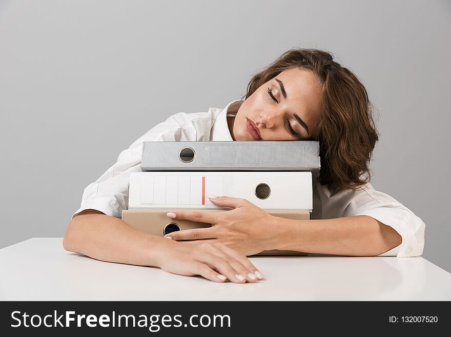 Photo of a bored tired young woman sitting at the table isolated over grey background sleeping on folders