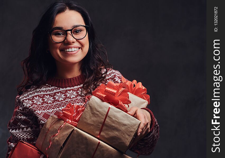 Portrait of a happy brunette girl wearing eyeglasses and warm sweater holding a gifts boxes, isolated on a dark textured