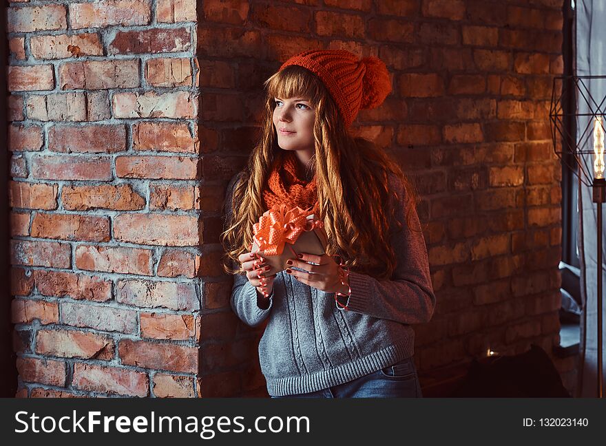 Christmas, Valentine`s Day, New year. Portrait of a lonely redhead girl wearing a warm sweater and hat holding a gift box while leaning on a brick wall.