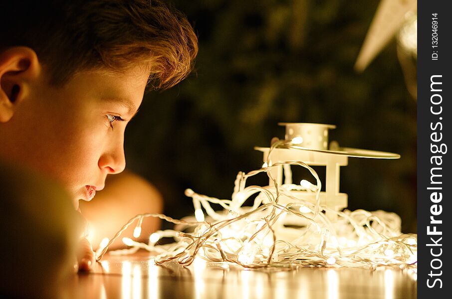 Cute young boy in white t shirt looks at lights in the evening at home in front of fir tree with lights.