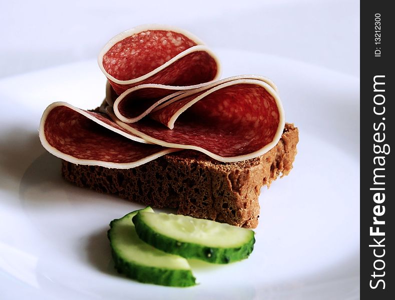 Few pieces of salami on a grey bread and two pieces of cucumber in the front. Few pieces of salami on a grey bread and two pieces of cucumber in the front.