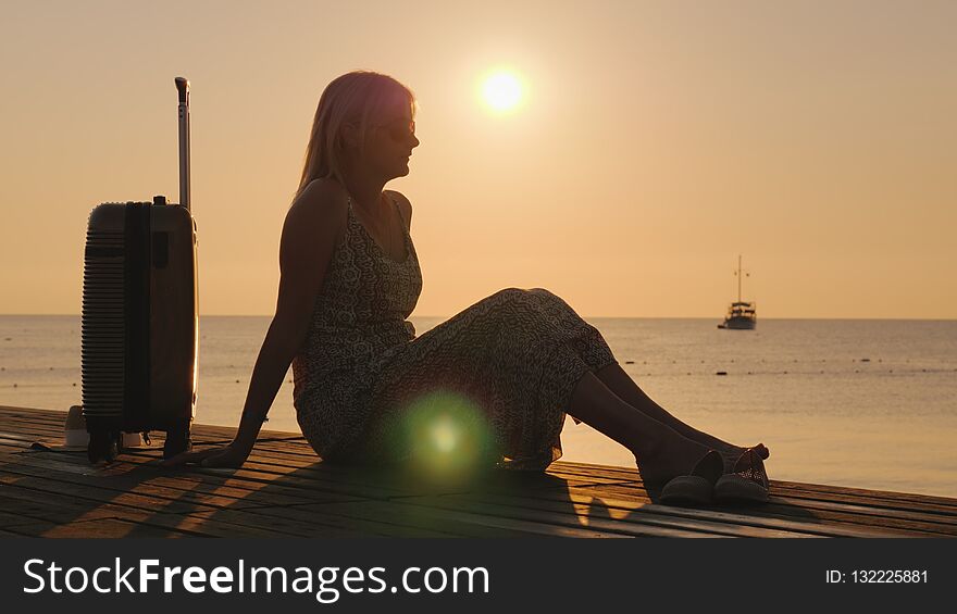Romantic woman with luggage looking at the ship far into the sea. At sunrise