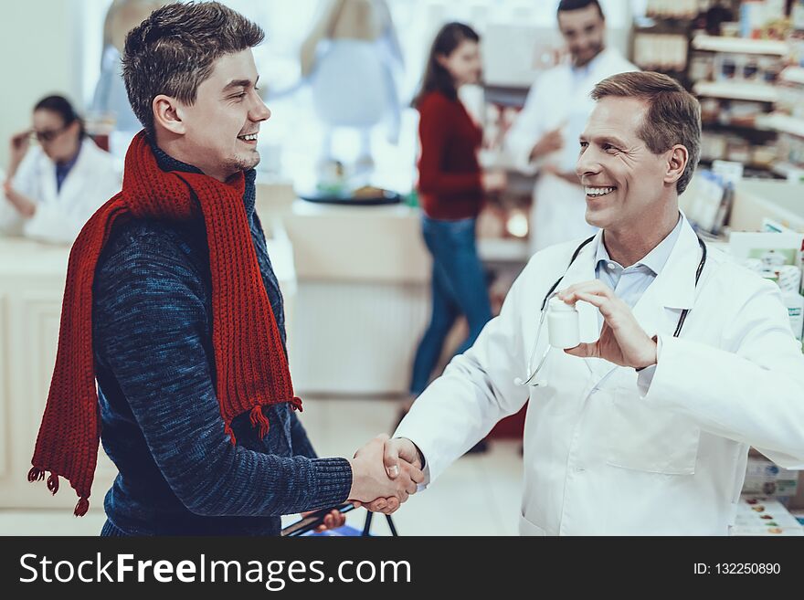 Smiling Pharmacist Handshaking with Customer. Pharmacist Showing a Bottle with Pills to Client. Other Pharmacists and Female Customer on Background. Pharmacists in Medical Uniform. People in Pharmacy.