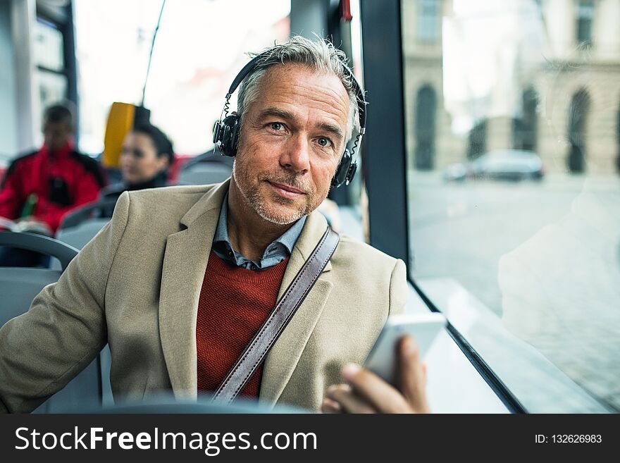 Mature tired businessman with heaphones and smartpone travelling by bus in city.