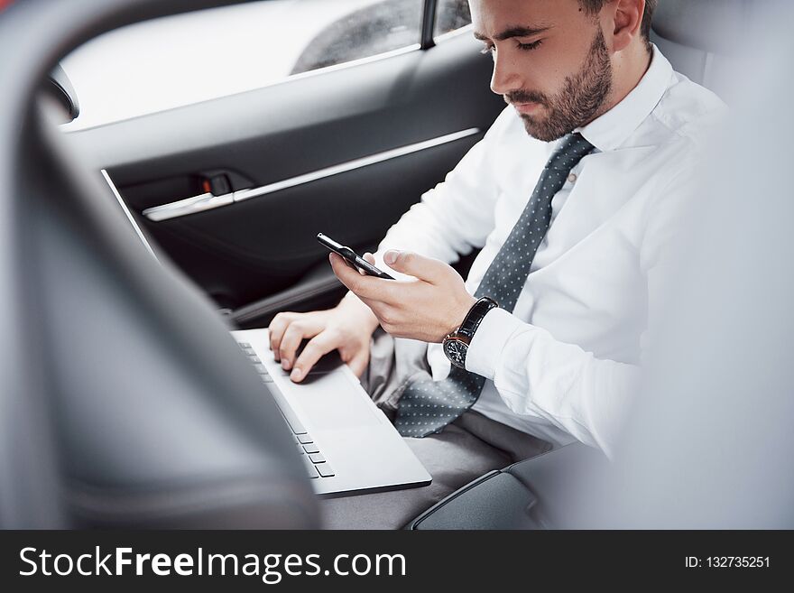 A young businessman working on laptop and talking on the phone while sitting in the car`s back. Works in motion
