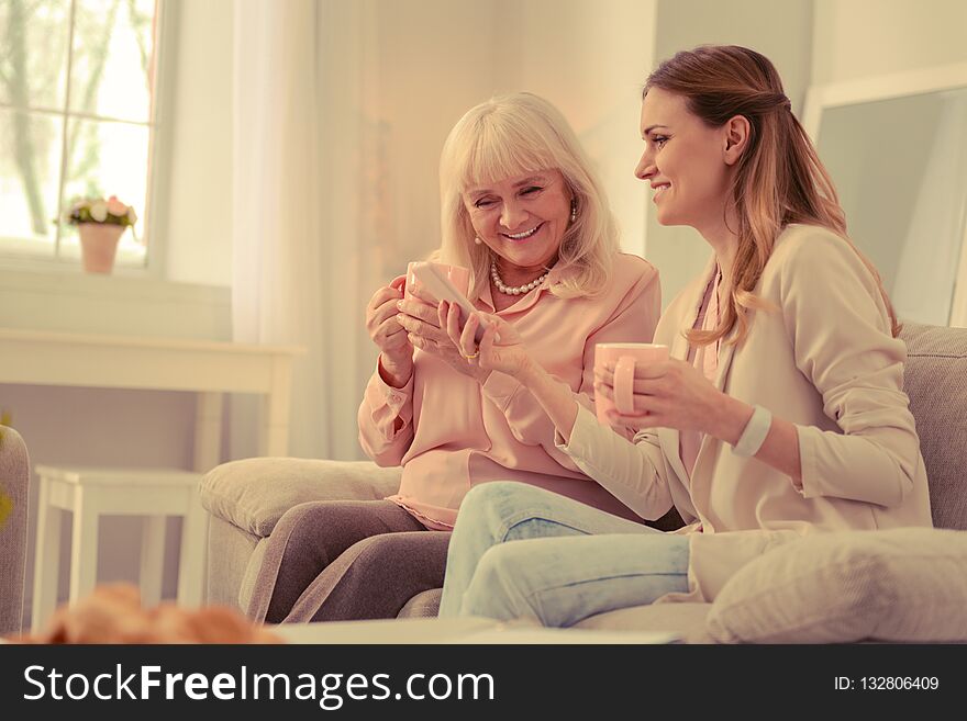 New smartphone. Happy cheerful women using a modern smartphone while sitting together on the sofa. New smartphone. Happy cheerful women using a modern smartphone while sitting together on the sofa