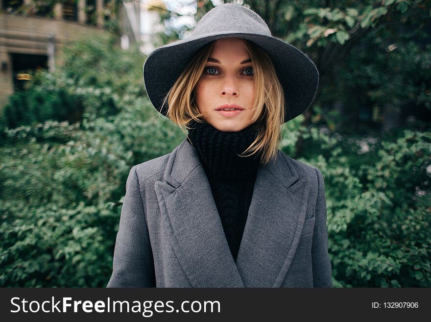 Photo of young blonde in gray coat on blurred background of city, green trees in afternoon