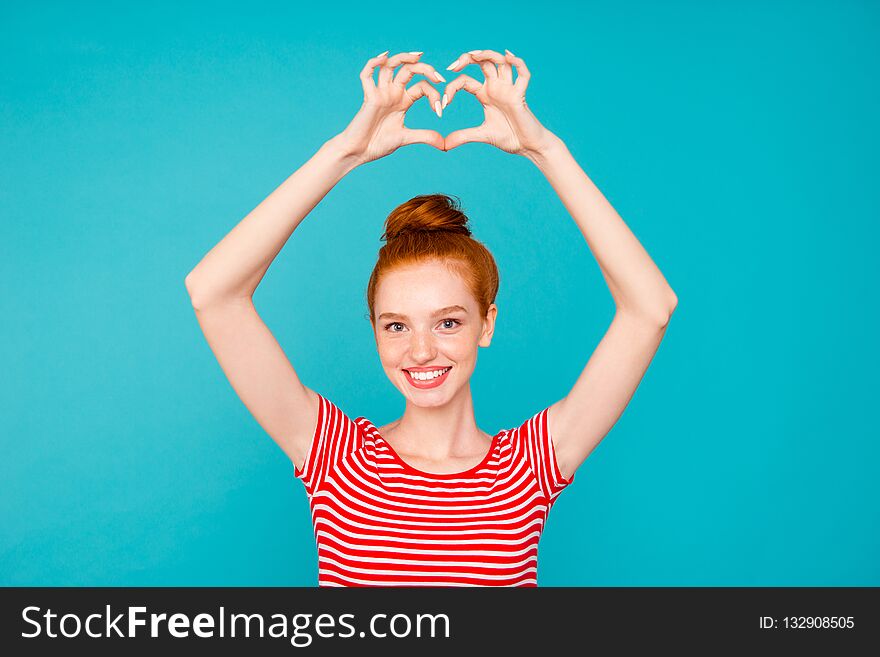 Portrait of nice cute adorable attractive lovely stylish cheerful positive red-haired girl with bun, wearing t-shirt, raising hands up, showing heart, isolated on bright vivid blue background