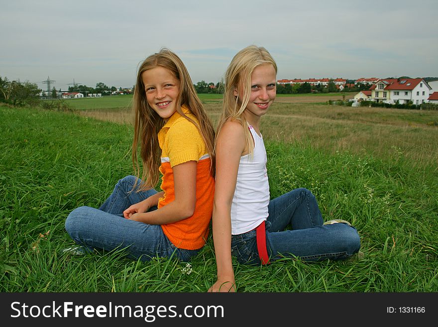 Two younf girl friends sitting in cross-legged pose in field. Two younf girl friends sitting in cross-legged pose in field
