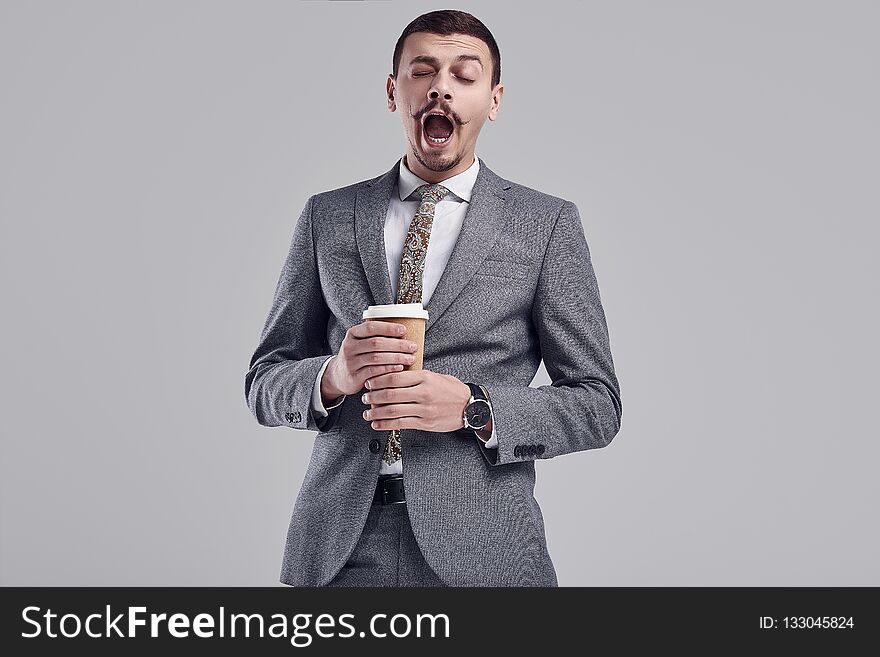 Portrait of handsome young confident arabic businessman with fancy mustache in fashion gray full suit yawns with a cup of coffee on studio background. Portrait of handsome young confident arabic businessman with fancy mustache in fashion gray full suit yawns with a cup of coffee on studio background