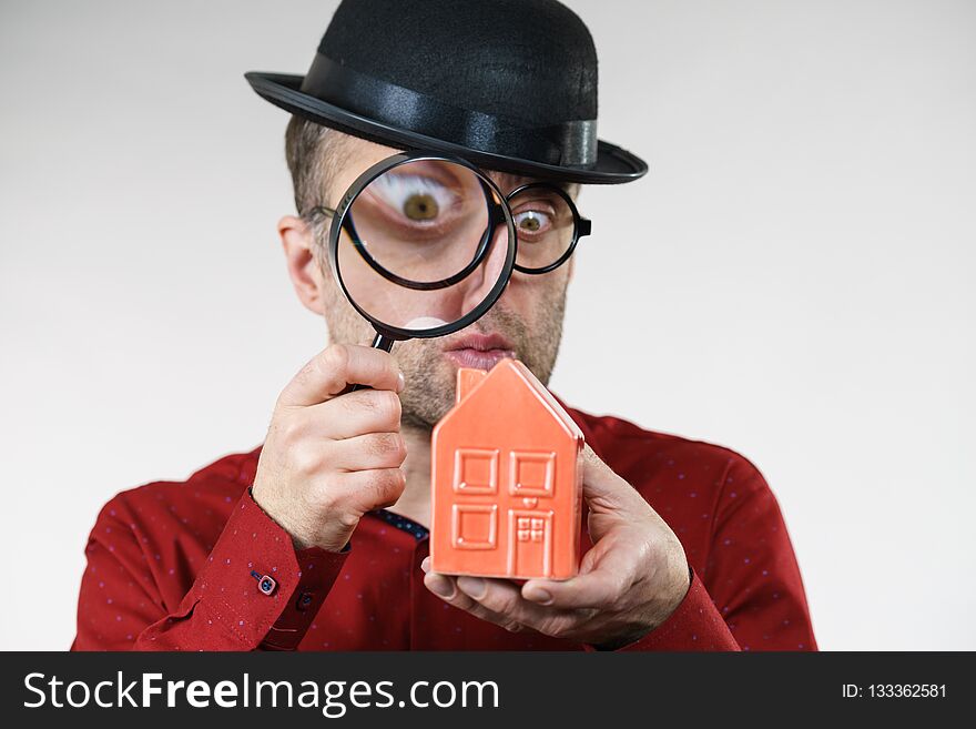 Funny looking adult man examining red model home using a magnifying glass, house inspection and real estate concept. Funny looking adult man examining red model home using a magnifying glass, house inspection and real estate concept