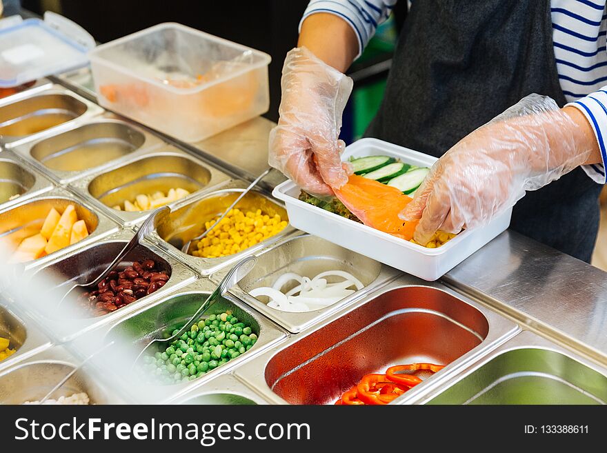 Fresh Salad bar counter with person`s hands lifting salmon into a salad box for healthy and diet meal with smooth light and shadow