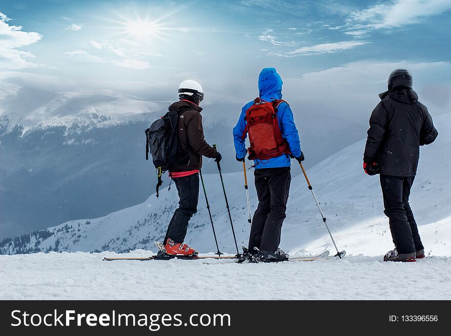 Three people in ski suits are sitting on the snow at the top of the mountain and looking into the distance. Family of skiers resting on the top of the mountain. back view