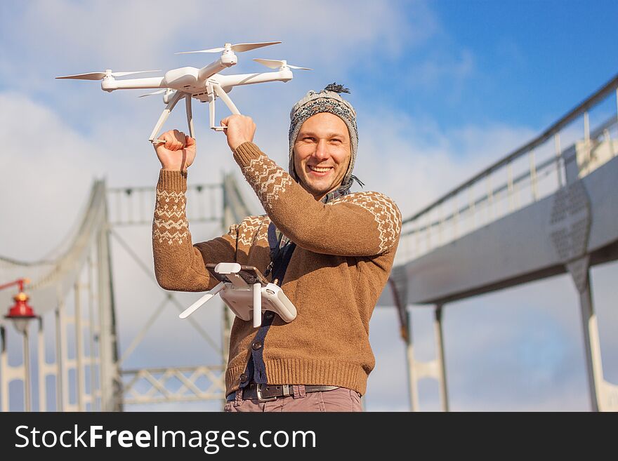 A young handsome man of European appearance launches a drone in the background of a blue sky on a bridge in winter. Travel man holding drone quad copter and remote control.