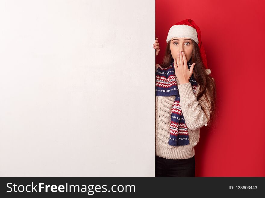 Young woman wearing sweater scarf and santa hat studio standing isolated on red wall holding white board looking camera covering mouth shocked. Young woman wearing sweater scarf and santa hat studio standing isolated on red wall holding white board looking camera covering mouth shocked
