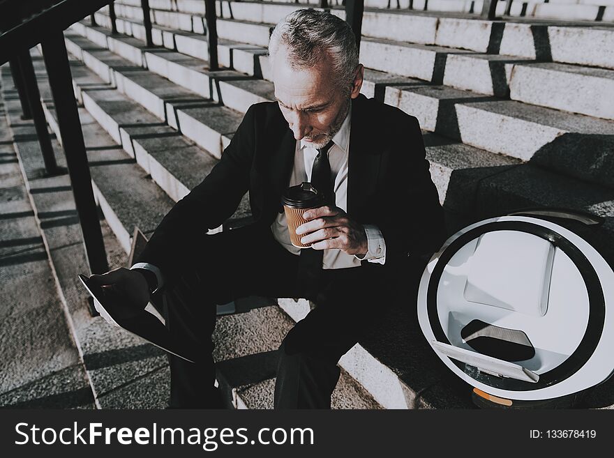 Businessman is Using a Tablet PC. Businessman is Old Smiling Man. Man is Holding a Coffee Cup. Man Wearing in Black Suit. Businessman is Sitting on Staircase. Monowheel near a Man. Sunny Daytime