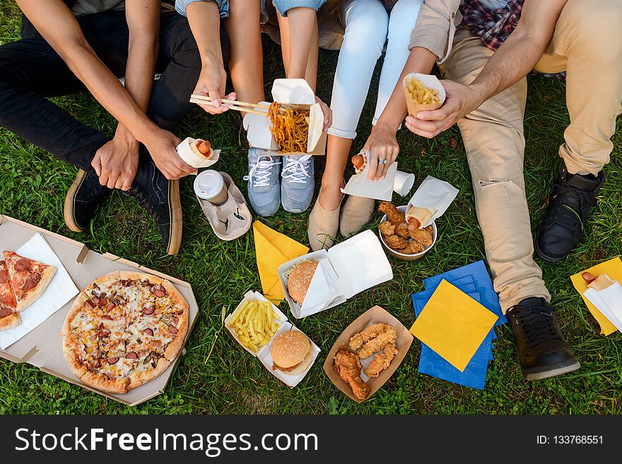 Junk food and bad eating habits concept. Friends sitting on the grass and eating, top view.