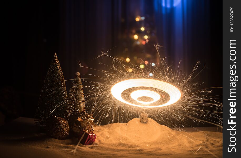 Glittering burning sparkler on snow with blurred Christmas tree on dark background. New Year Holiday concept with empty space for your text. Selective focus