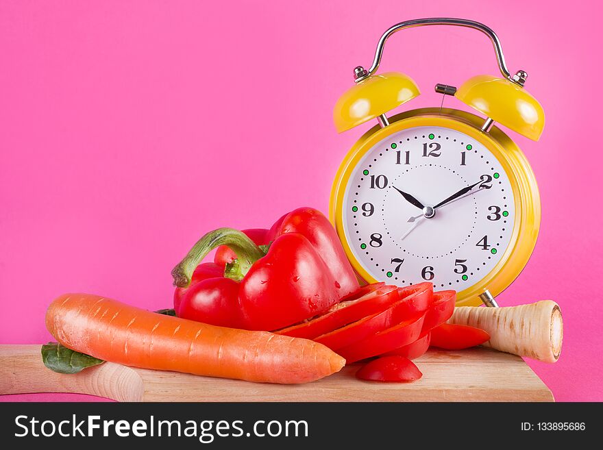 Fresh vegetables on a wooden kitchen board and retro clock isolated on pink background. Red pepper, carrots, parsnip and salad on plate. Chrono diet and nutrition concept. Close up, selective focus