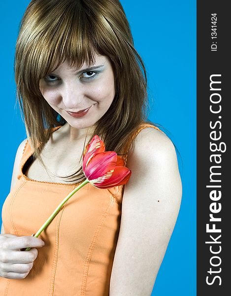 Portrait of a young girl smiling, with a red tulip in her hand, looking confident in her youth and looks; nice make-up; posing in a studio; expression of youth; isolated on blue background. Portrait of a young girl smiling, with a red tulip in her hand, looking confident in her youth and looks; nice make-up; posing in a studio; expression of youth; isolated on blue background