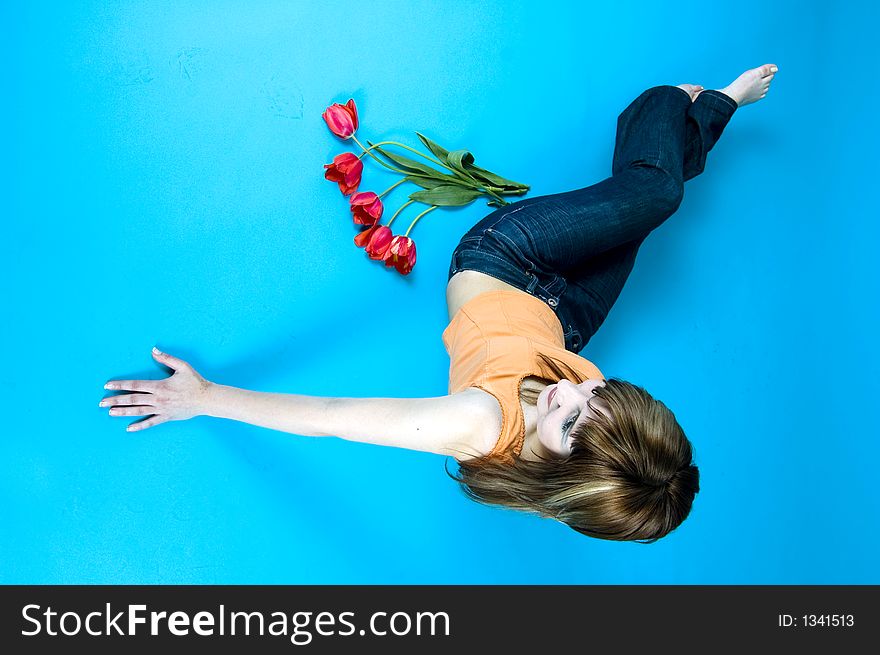 Portrait of a young girl laying on the floor and looking at a bouquet of red tulips and smiling, expression of youth and happiness; nice make-up, isolated on blue background. Portrait of a young girl laying on the floor and looking at a bouquet of red tulips and smiling, expression of youth and happiness; nice make-up, isolated on blue background