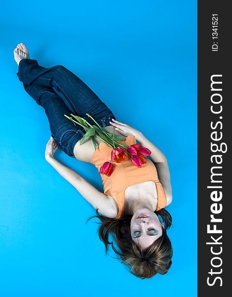 Portrait of a young girl laying on the floor with a bouquet of tulips covering her chest, expression of youth and senses; nice make-up, isolated on blue background. Portrait of a young girl laying on the floor with a bouquet of tulips covering her chest, expression of youth and senses; nice make-up, isolated on blue background