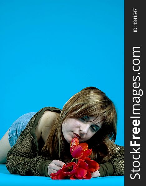 Portrait of a young girl laying on the floor and looking at a bouquet of red tulips, expression of youth and senses; nice make-up, isolated on blue background. Portrait of a young girl laying on the floor and looking at a bouquet of red tulips, expression of youth and senses; nice make-up, isolated on blue background