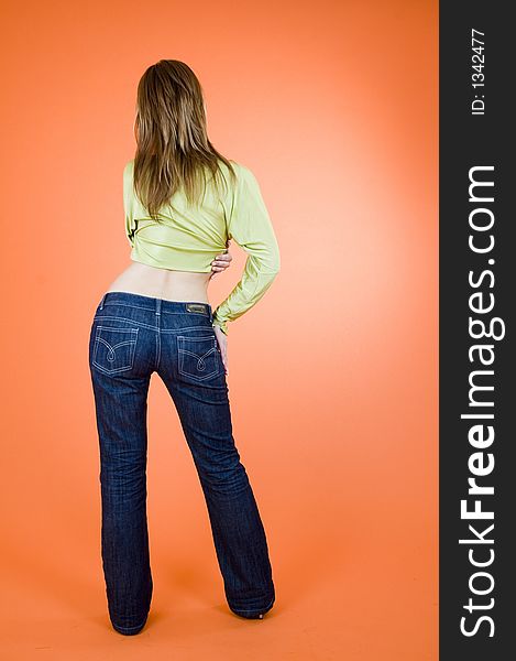Blond girl wearing jeans standing with her back at the viewer; posing in a studio; orange background. Blond girl wearing jeans standing with her back at the viewer; posing in a studio; orange background