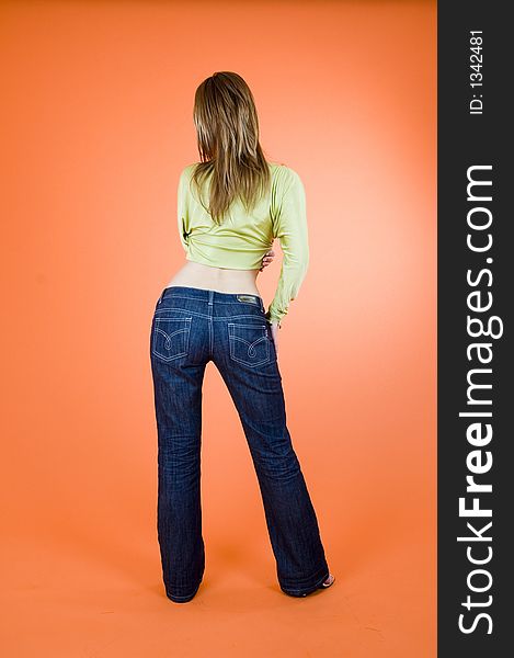 Blond girl wearing jeans standing with her back at the viewer; posing in a studio; orange background. Blond girl wearing jeans standing with her back at the viewer; posing in a studio; orange background