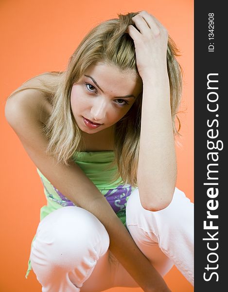 Blond girl relaxing on the floor and playing with a hand in her hair; wearing casual clothes; orange background. Blond girl relaxing on the floor and playing with a hand in her hair; wearing casual clothes; orange background