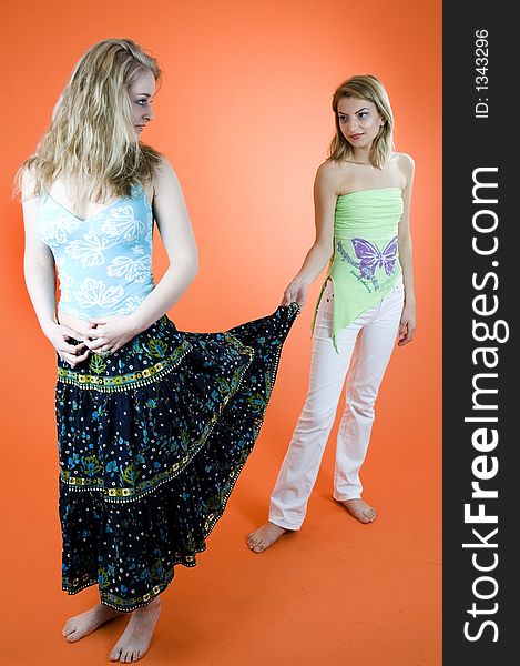 Two beautiful blond girls, one pulling the skirt of another, smiling and looking at her; orange background. Two beautiful blond girls, one pulling the skirt of another, smiling and looking at her; orange background