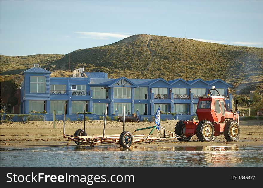 Tractor pulling a whelled structure on beachfront in front of a blue building. Tractor pulling a whelled structure on beachfront in front of a blue building