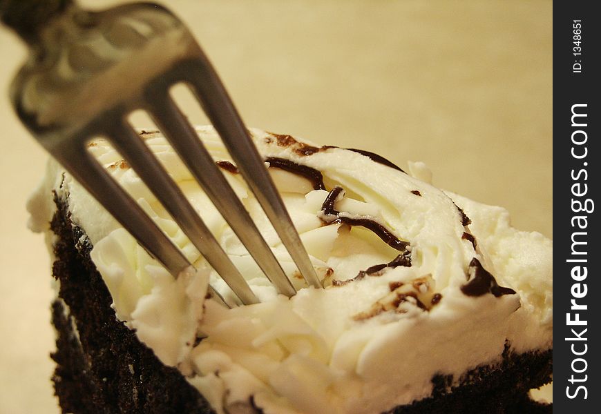 A large wedge of chocolate cake with white icing and white chocolate shavings on top with a fork digging in for a bite. A large wedge of chocolate cake with white icing and white chocolate shavings on top with a fork digging in for a bite.