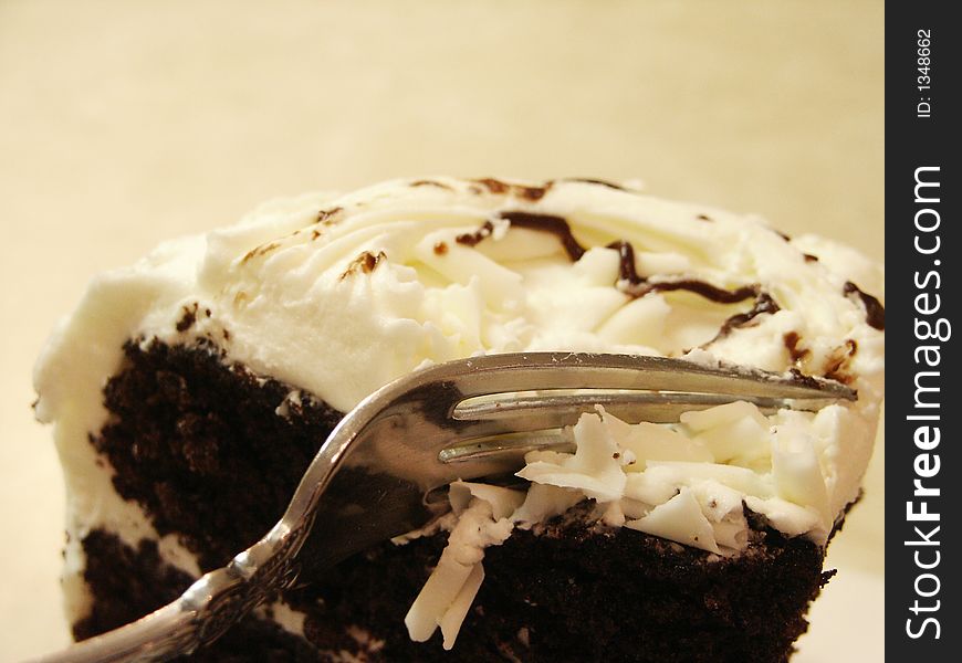 A large wedge of chocolate cake with white icing and white chocolate shavings on top with a fork digging in for a bite. A large wedge of chocolate cake with white icing and white chocolate shavings on top with a fork digging in for a bite.