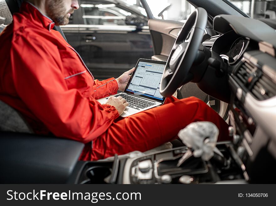 Auto mechanic in red uniform diagnosing car with computer sitting on the driver seat at the car service. Auto mechanic in red uniform diagnosing car with computer sitting on the driver seat at the car service