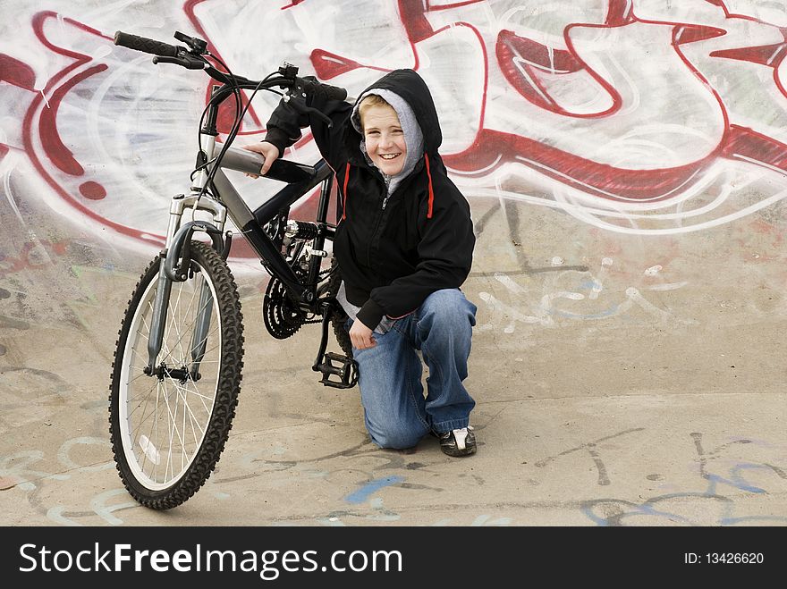 A horizontal image of a young teenager with a cheeky smile kneeling beside his bicycle in a graffeti covered skatepark bowl