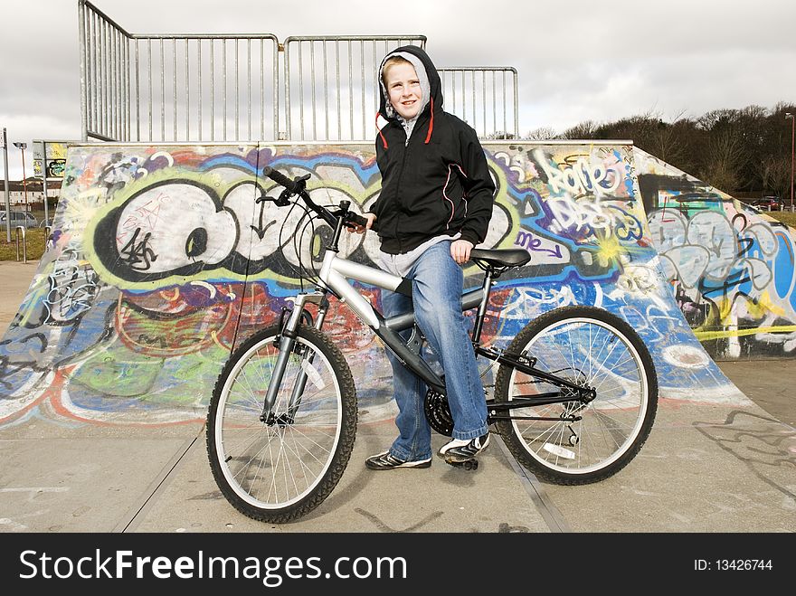 A horizontal image of a male teenager astride his bicycle in front of a skatepark ramp. A horizontal image of a male teenager astride his bicycle in front of a skatepark ramp