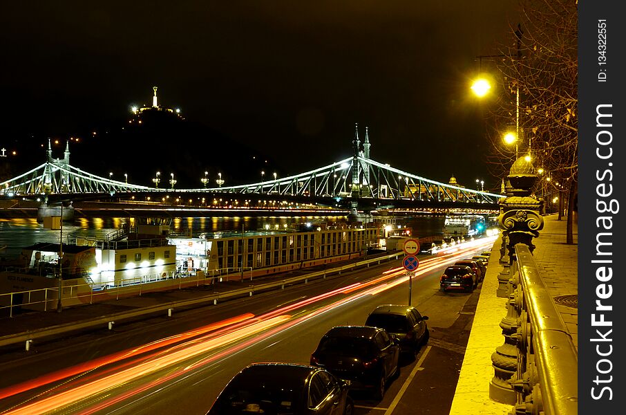 brightly lit night panorama of the Liberty bridge in Budapest with street lighting and the Gellert hill above and parked cars along the well and brightly lit road with cast iron steel railing