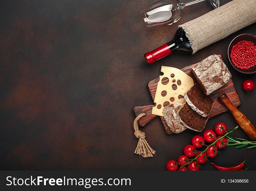 Maasdam cheese sliced on a cutting board with cherry tomatoes, black bread, garlic and rosemary, bottle of wine, wine glass on rusty background. Top view