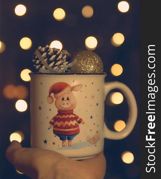 Christmas theme. in the hand of a mug with a pig and Christmas balls. bokeh in the background. Twinkling lights on the holiday