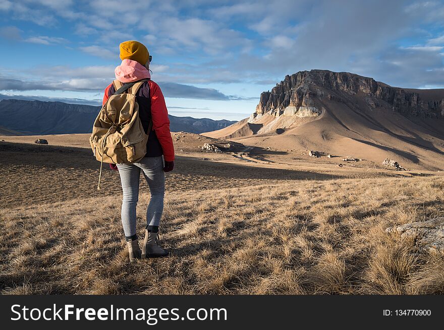 Portrait from the back of a girl traveler in a jacket with a cap and a backpack stands on the background of an epic