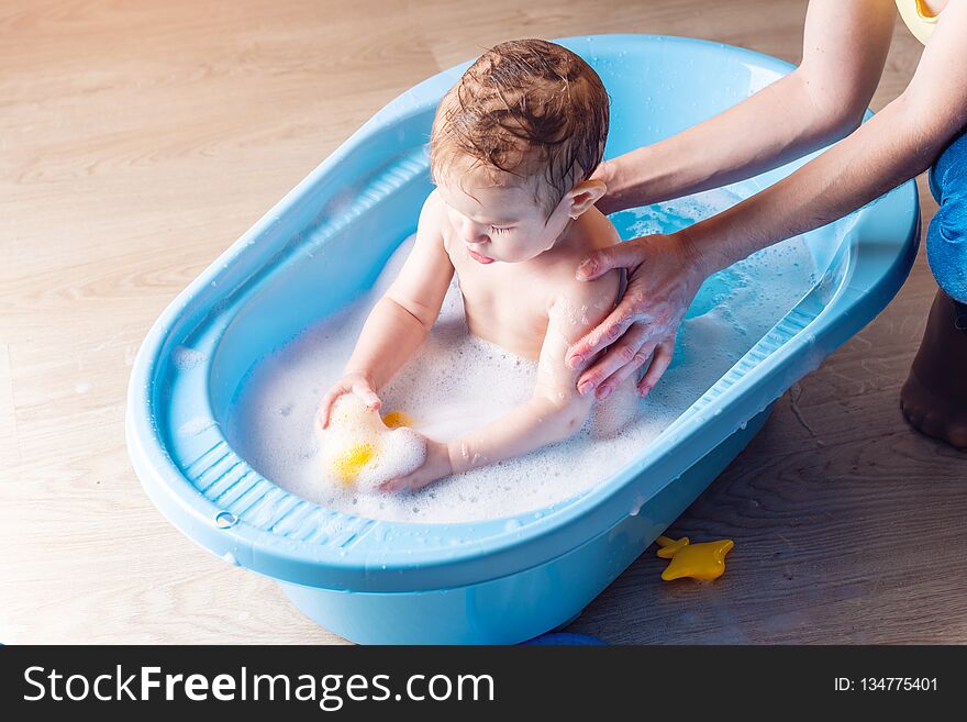 Mom washing little boy in a blue bath in the bathroom. Baby playing with a yellow duck and soap bubbles