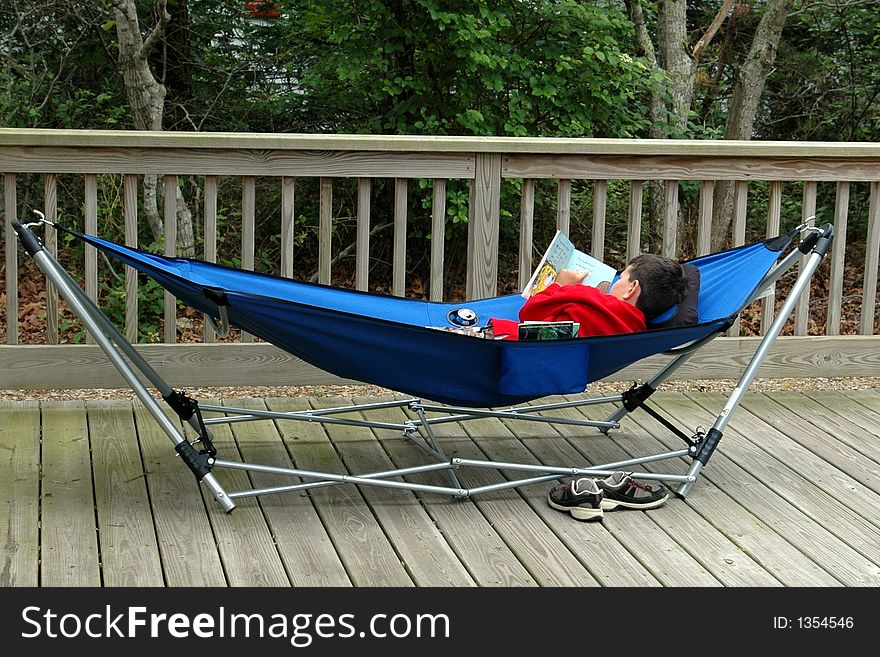 A young boy is laying in a blue hammock reading a book. A young boy is laying in a blue hammock reading a book.