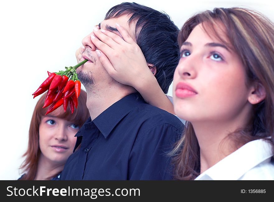 Guy holding a bunch of red chilli peppers in his mouth and two beautiful girls covering his eyes with the hands. Guy holding a bunch of red chilli peppers in his mouth and two beautiful girls covering his eyes with the hands