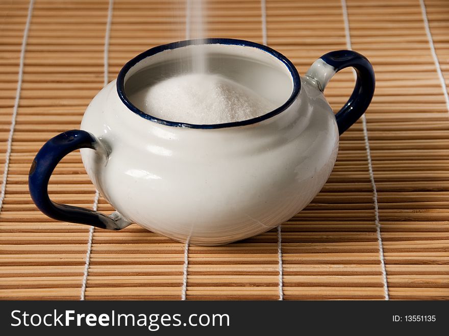 A white enamel sugar bowl on a straw mat being filled with sugar. A white enamel sugar bowl on a straw mat being filled with sugar
